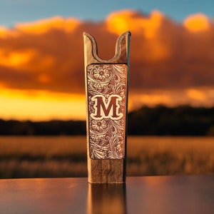 May include: A wooden boot jack with a decorative floral design and the letter "M" in the center. The boot jack is made of dark brown wood and has a curved top for pulling off boots. The background features a sunset.