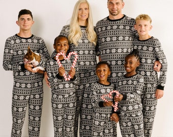 Può includere: Una famiglia di sette persone che indossa pigiami abbinati a motivi neri e bianchi con un design a fiocco di neve. I pigiami hanno maniche lunghe e pantaloni. La famiglia è in piedi davanti a un muro bianco.