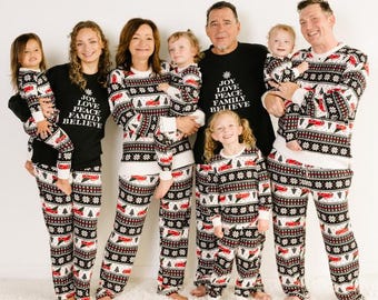 May include: A family of seven wearing matching Christmas pajamas with a red truck and snowflake pattern. The pajamas are white with black and red accents. The adults are wearing black shirts with the words "Joy Love Peace Family Believe" printed on them.