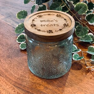 May include: A glass jar with a wooden lid. The lid is engraved with the text "Millie's treats" and paw prints. The jar has a textured surface.