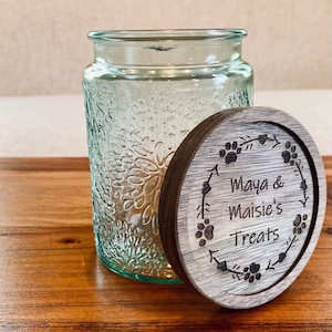 May include: A clear glass jar with a textured surface and a wooden lid. The lid has a design of paw prints and a wreath with the text "Maya & Maisie's Treats".