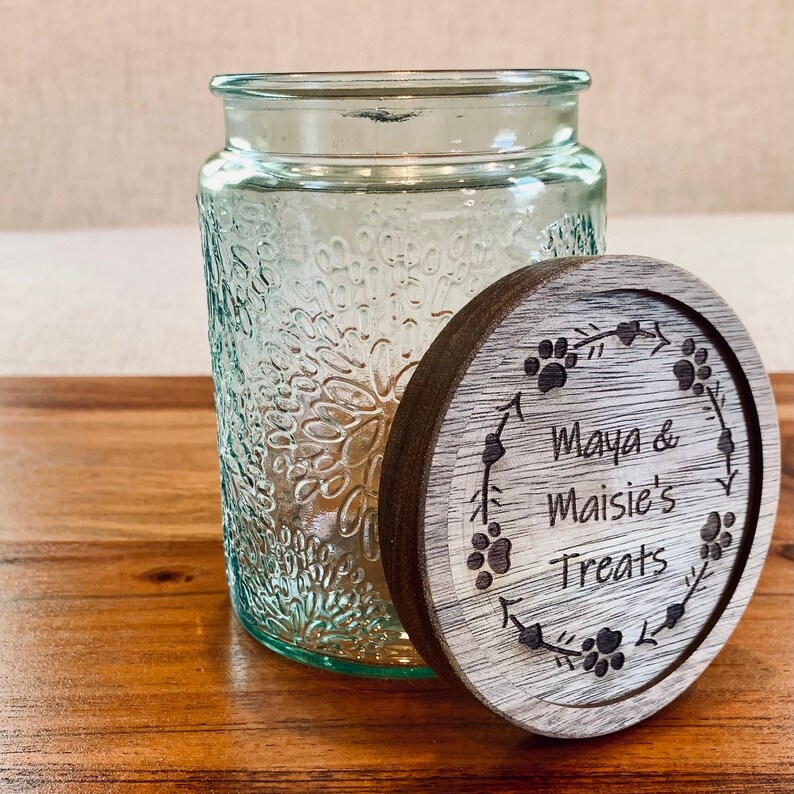 May include: A clear glass jar with a textured surface and a wooden lid. The lid has a design of paw prints and a wreath with the text "Maya & Maisie's Treats".