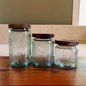 May include: Three glass canisters with wooden lids. The canisters are clear glass with a textured, floral pattern. The lids are a dark brown wood.