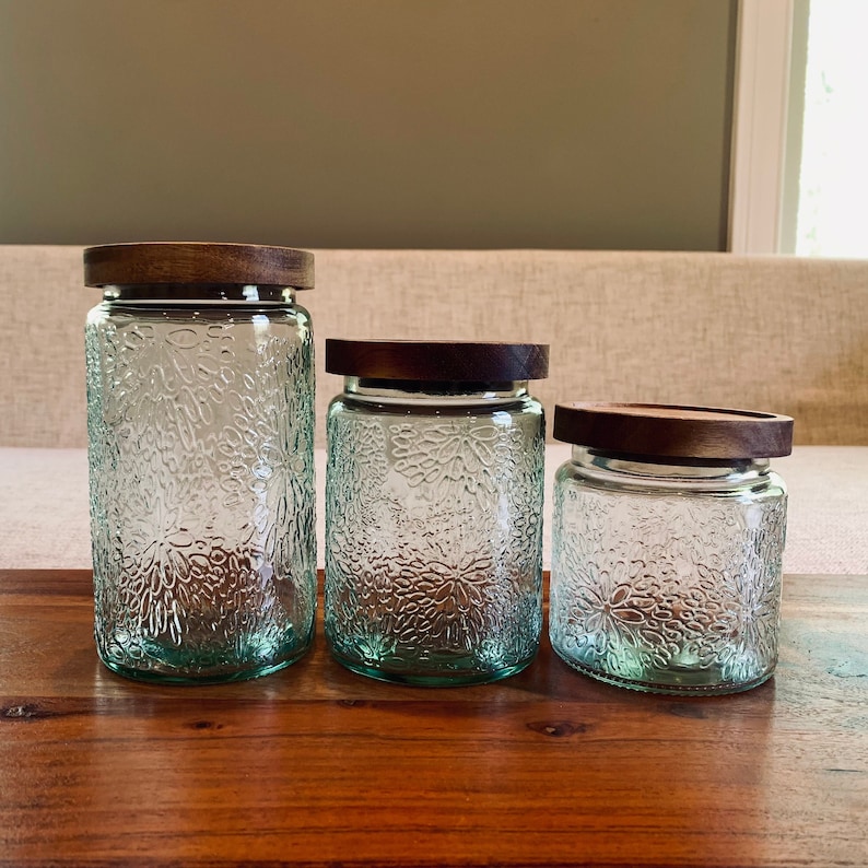 May include: Three glass canisters with wooden lids. The canisters are clear glass with a textured, floral pattern. The lids are a dark brown wood.