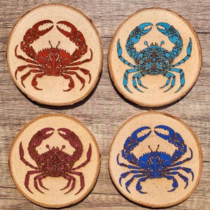 May include: Four round wooden coasters with a decorative crab design. Two coasters feature a blue crab with intricate details, and two coasters feature a red crab with intricate details.