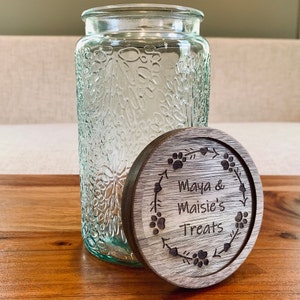 May include: A clear glass jar with a textured surface and a wooden lid. The lid has a design of paw prints and a wreath with the text "Maya & Maisie's Treats".