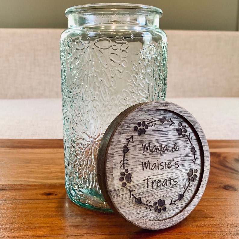 May include: A clear glass jar with a textured surface and a wooden lid. The lid has a design of paw prints and a wreath with the text "Maya & Maisie's Treats".