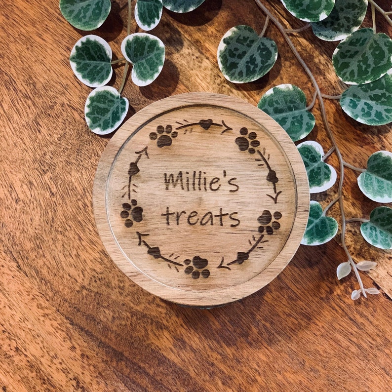 May include: A wooden round treat jar with the text "Millie's treats" engraved on it. The jar is decorated with a paw print and arrow design.