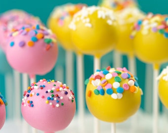 Puede incluir: Primer plano de cake pops decorados con chispas de colores. Los cake pops están en palos y dispuestos en una fila. Algunos son amarillos y otros son rosados.