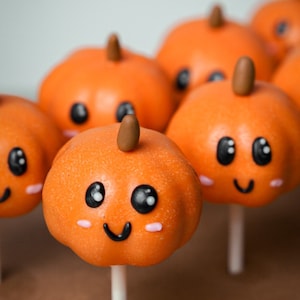 May include: A close-up of a group of orange pumpkin-shaped cake pops with cute cartoon faces. The cake pops are on sticks and arranged on a brown surface.