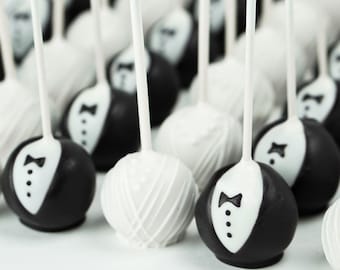 May include: A collection of black and white cake pops decorated with tuxedo and wedding dress designs. The cake pops are on sticks and arranged in a row.