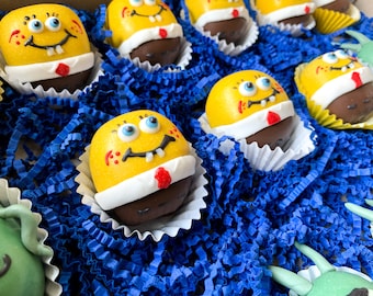 May include: A box of yellow and brown chocolate-covered treats decorated with cartoon faces. The treats are arranged in a blue shredded paper.
