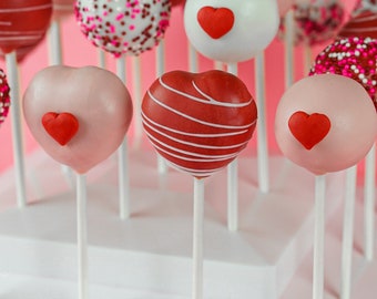 May include: A tiered white cake stand with heart-shaped cake pops decorated with red, pink, and white frosting. Some cake pops have red heart sprinkles and others have white sprinkles.