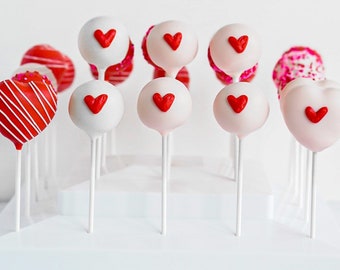 May include: A white cake pop stand with red and white cake pops decorated with red hearts. The cake pops are arranged in rows on the stand.
