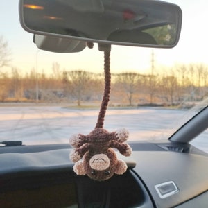 Può includere: Una scimmia di uncinetto marrone appesa allo specchietto retrovisore di un'auto. La scimmia ha un viso marrone chiaro e una pancia marrone chiaro.
