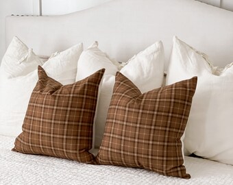 May include: Two brown and beige plaid throw pillows on a white bed with white linen pillows. The bed has a white quilted bedspread and a beige throw blanket.