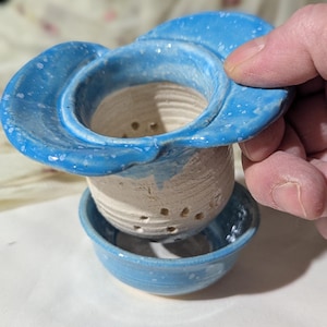 May include: A handmade ceramic tea infuser set. The set includes a light blue, flower-shaped lid, a beige infuser with drainage holes, and a small blue saucer. The infuser is held by a hand.