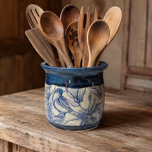 May include: A blue and cream ceramic utensil holder with a bird and floral design, filled with various wooden kitchen utensils. The holder sits on a wooden surface, showcasing a rustic aesthetic.