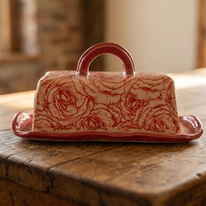 May include: A rectangular ceramic butter dish with a lid featuring a red rose pattern on a cream background. The lid has a curved red handle, and the base is a matching red tray with slightly raised edges, designed to hold a standard stick of butter.