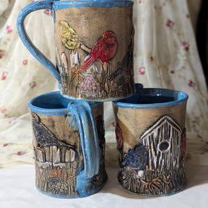May include: Three ceramic mugs with blue handles and rims. The mugs feature detailed relief designs of birds, birdhouses, and fences. The mugs have a brown background with colorful birds and floral accents.