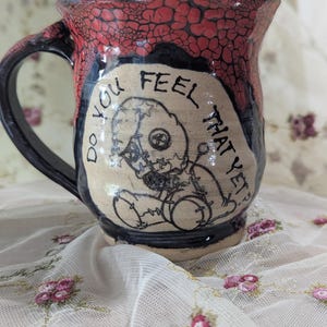 May include: A ceramic mug with a black handle and a red crackle glaze on the upper portion. The center features a beige panel with a black line drawing of a stitched doll and the text "DO YOU FEEL THAT YET?" written in black ink.