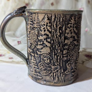 May include: A ceramic mug with a forest scene carved into the exterior. The mug features a detailed relief of trees, creatures, and a textured handle. The color palette includes shades of brown, black, and green.