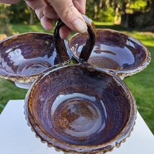 May include: A brown and purple ceramic serving dish with three bowls and a handle. The bowls have a scalloped edge and a glossy finish.