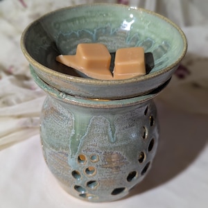 May include: A handcrafted ceramic wax warmer in shades of green and brown. The warmer has a bowl-shaped top with two wax cubes and a perforated base. The design features a rustic, glazed finish, perfect for home fragrance.