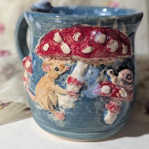 May include: A blue ceramic mug with a whimsical design featuring a red mushroom with white spots, a small mouse, and other mushrooms. The mug has a handle and is decorated with a light blue glaze.