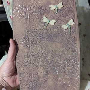 May include: A rectangular, lavender-colored cutting board with a handle. The board features an embossed, swirling pattern and is adorned with three dragonfly embellishments. The dragonflies are white with light blue accents. The board is held by a person.