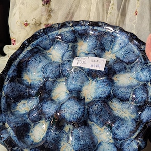 May include: Round ceramic plate with a textured surface. The plate features a pattern of blue and white organic shapes. The rim is dark blue and scalloped. The plate has a glossy finish.