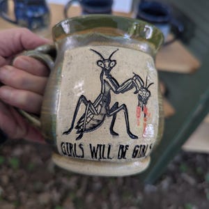 May include: A ceramic mug with a green and beige glaze featuring a black ink illustration of a praying mantis holding another insect with red accents. The text "GIRLS WILL BE GIRLS" is etched in black lettering at the bottom of the mug.