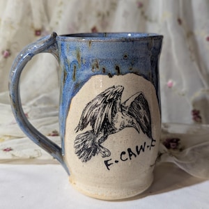May include: Handmade ceramic mug with a blue glazed top and handle. The mug features a black and white illustration of a bird with the text "F.CAW-" on a cream-colored background. The mug is suitable for hot or cold beverages.