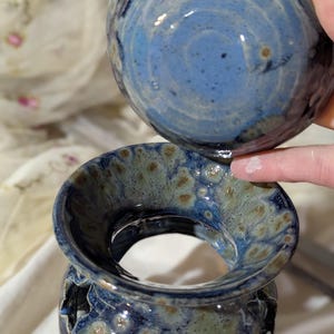 K&ouml;nnte beinhalten: Ein zweiteiliger Keramik-Duftlampenhalter mit blau-gr&uuml;ner Glasur. Das Oberteil ist eine runde Schale, und die Basis hat sternf&ouml;rmige Aussparungen. Der Halter wird von einer Hand gehalten.