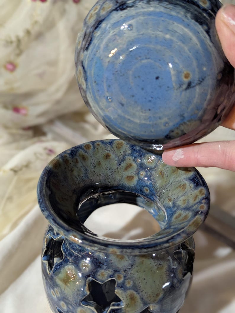 K&ouml;nnte beinhalten: Ein zweiteiliger Keramik-Duftlampenhalter mit blau-gr&uuml;ner Glasur. Das Oberteil ist eine runde Schale, und die Basis hat sternf&ouml;rmige Aussparungen. Der Halter wird von einer Hand gehalten.