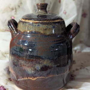 May include: A handmade ceramic jar with a lid, featuring a rich blend of brown, blue, and tan glazes. The jar has two loop handles and a small knob on the lid. The pottery has a glossy finish and a rustic, artisanal aesthetic.