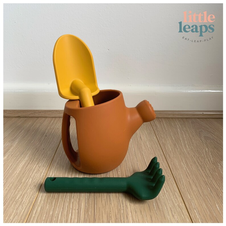 Pu&ograve; includere: Un annaffiatoio color terracotta con una pala gialla all'interno e un rastrello verde su una superficie di legno. L'immagine include anche il testo "little leaps eat-leap-play".