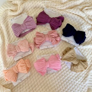 May include: Assortment of ten fabric hair bows in various shades of pink, beige, and brown. The bows are arranged on a white knitted surface. Each bow is tied with a knot in the centre and has a slightly ruffled texture.