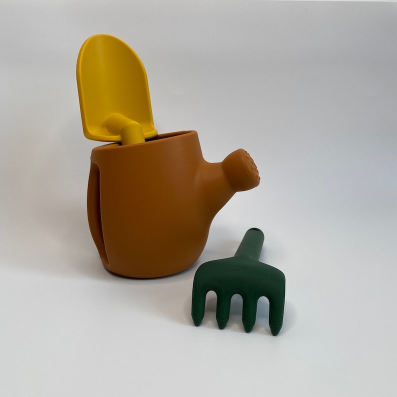 Pu&ograve; includere: Un annaffiatoio marrone con una pala gialla e un rastrello verde scuro. L'annaffiatoio ha un beccuccio e un manico. La pala &egrave; inserita nella parte superiore dell'annaffiatoio. Il rastrello ha quattro denti.
