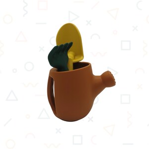 Pu&ograve; includere: Un annaffiatoio marrone con manico e beccuccio, contenente una cazzuola gialla e un guanto da giardinaggio verde. Gli attrezzi sono disposti all'interno dell'annaffiatoio, creando una scena giocosa.