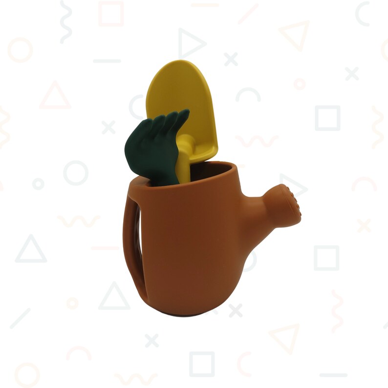 Pu&ograve; includere: Un annaffiatoio marrone con manico e beccuccio, contenente una cazzuola gialla e un guanto da giardinaggio verde. Gli attrezzi sono disposti all'interno dell'annaffiatoio, creando una scena giocosa.