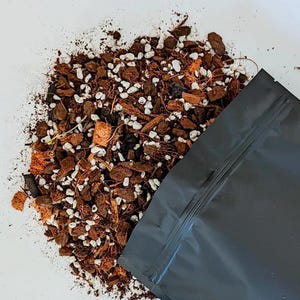 May include: A black plastic bag with a zipper closure partially filled with a brown and white soil mix. The soil mix contains small pieces of bark, coconut husk, and white pebbles.