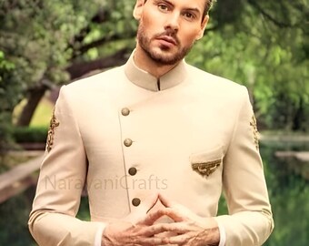 Pode incluir: Um blazer bege com botões e bordados dourados. O blazer tem uma longa echarpe fluida presa nas costas. O modelo está usando calças marrons.