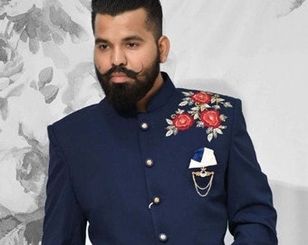 Puede incluir: Una chaqueta bandhgala azul marino con cuello mao, botones plateados y bordado floral rojo en el hombro izquierdo. La chaqueta incluye un pañuelo de bolsillo blanco con un broche de cadena dorado, combinado con pantalones blancos.