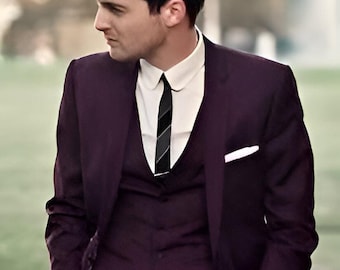 Pode incluir: Um homem vestindo um terno roxo escuro de três peças com uma camisa branca de colarinho e uma gravata com padrão preto e branco. Ele está em pé em uma calçada de tijolos.