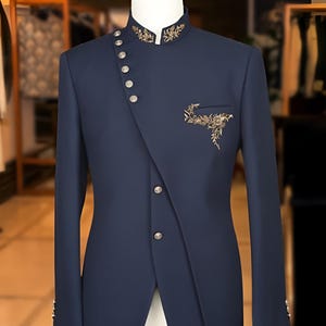 May include: Navy blue suit jacket with an asymmetrical button closure and mandarin collar. The jacket features gold embroidery on the collar and pocket, and silver buttons. The tailored fit and unique design make it a stylish formal wear option.