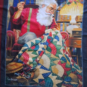 May include: A fabric panel depicting Santa Claus sewing a quilt. Santa wears a red shirt with green braces and glasses. The quilt has a colourful patchwork design with red, blue, green, and tan fabrics. A basket of sewing supplies is in the foreground. The artist's name, Tom Newsom, is visible.