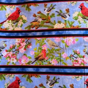 May include: A patterned fabric featuring birds, flowers, and foliage. The design includes red cardinals, grey and brown birds, pink blossoms, and green pine needles on a light blue backdrop. Black and red stripes border the design.