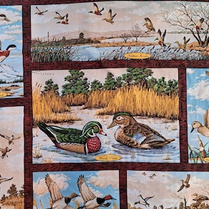 May include: A fabric panel with illustrations of ducks in various scenes. The artwork shows different duck species in flight and at rest, set against backgrounds of water, trees, and sky. The panel is framed with a dark brown border.