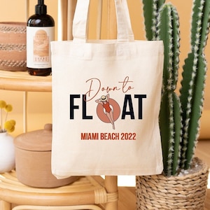 Puede incluir: Bolso tote de color crema con las palabras "Down to Float" y "Miami Beach 2022" impresas. El diseño incluye una persona en traje de baño en un flotador. El bolso cuelga de una estantería de bambú.